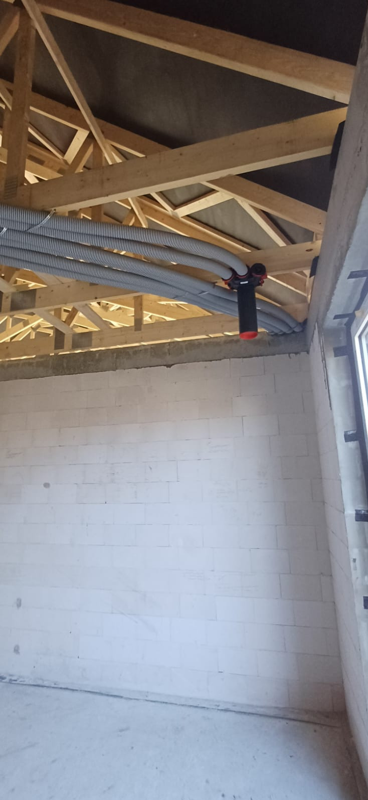 Szare, karbowane rury wentylacyjne pod drewnianą konstrukcją dachu, z elementami montażowymi w kolorze czarno-czerwonym. Surowy stan budynku, białe ściany.