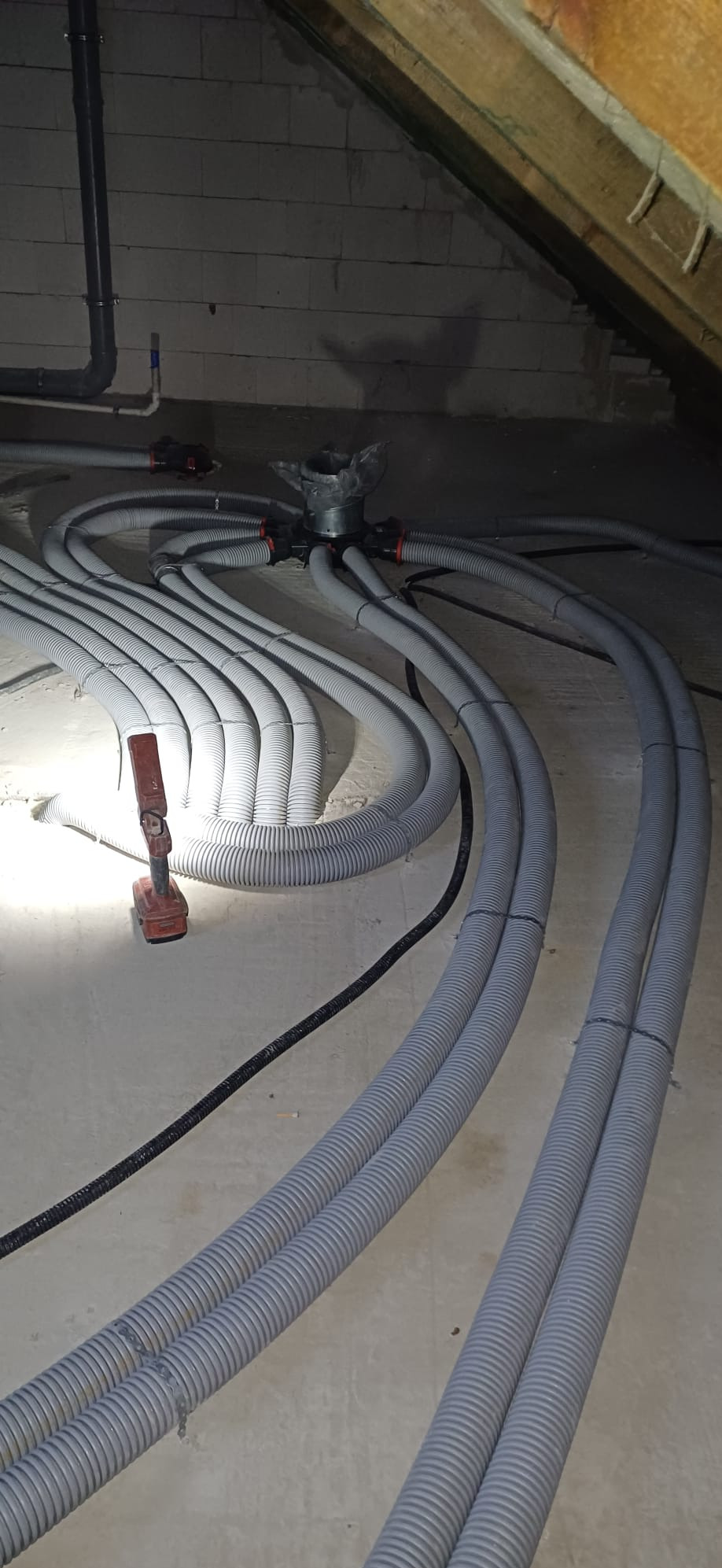 Szare, karbowane rury wentylacyjne rozchodzą się promieniście od metalowego rozdzielacza na poddaszu, obok leży wiertarka akumulatorowa.