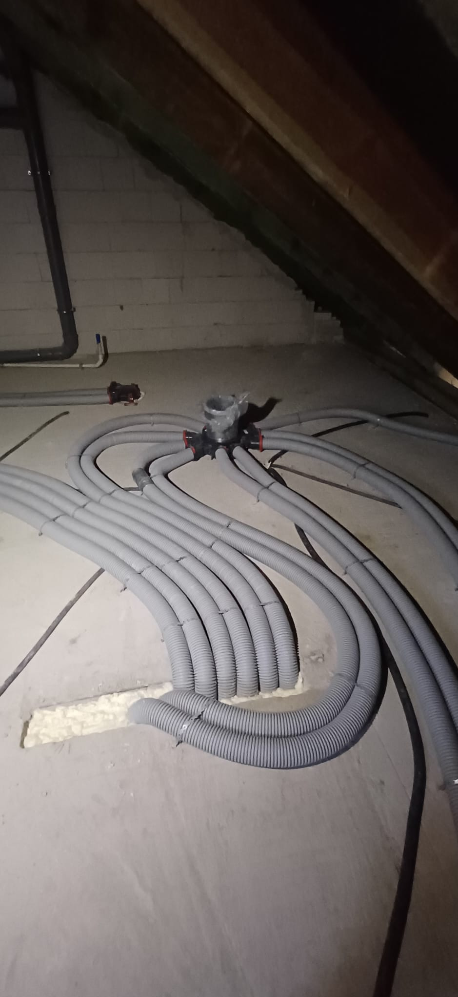 Szare, karbowane rury wentylacyjne rozchodzą się promieniście z czarnej rozdzielnicy na poddaszu. Fragmenty rur w ścianie uszczelnione pianką.