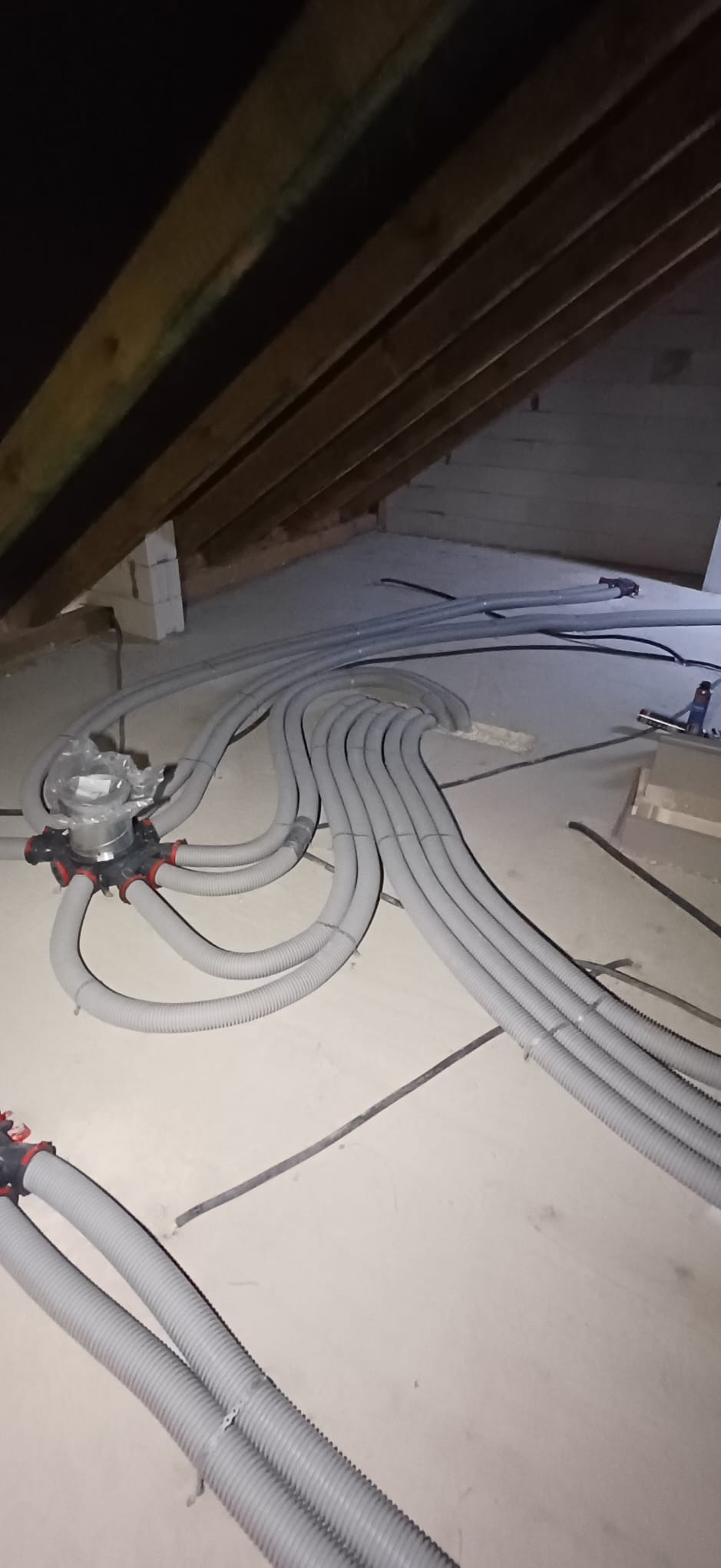 Szare, elastyczne rury wentylacyjne ułożone na poddaszu, prowadzące do rozdzielacza. Widoczne drewniane elementy konstrukcji dachu i betonowa posadzka.