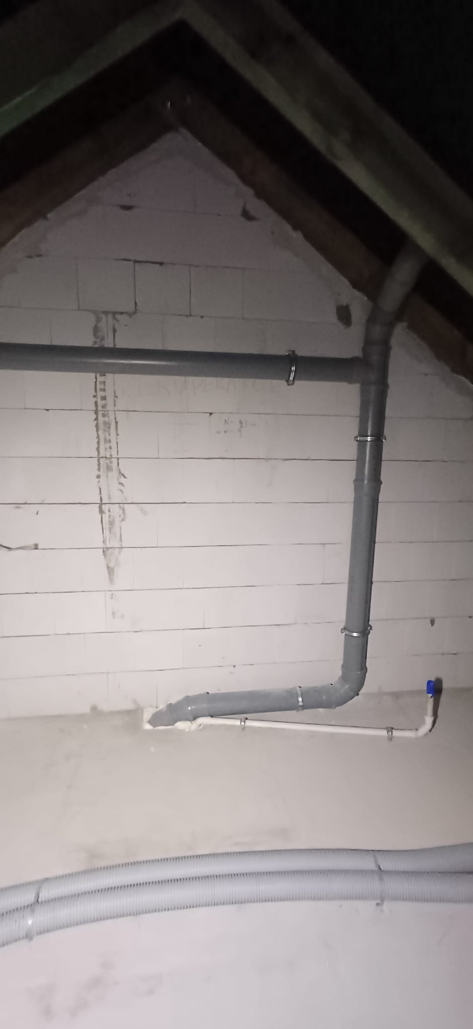 Szare rury kanalizacyjne zamocowane do ściany z białych bloczków w poddaszu, widoczne elementy instalacji sanitarnej i fragment instalacji elektrycznej w peszlu.
