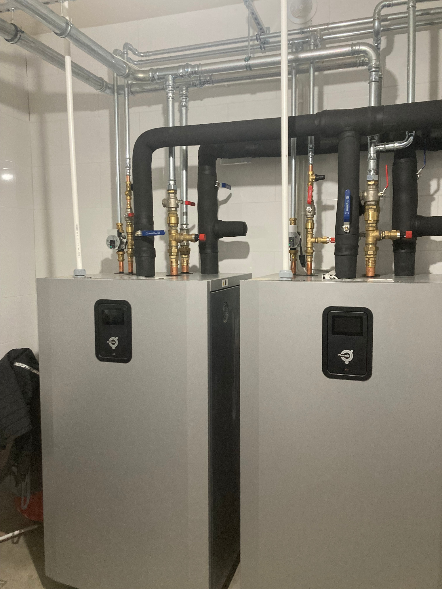 Dwie szare pompy ciepła Thermia z czarnymi izolowanymi rurami i zaworami na tle białych płytek. Instalacja grzewcza w kotłowni.