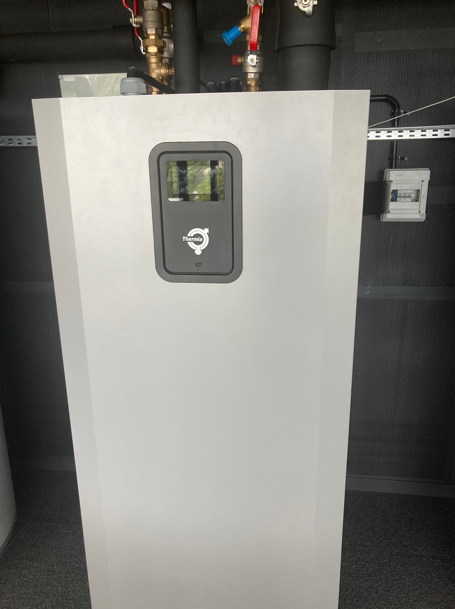 Biała pompa ciepła Thermia w kotłowni z szarą, perforowaną ścianą w tle, widoczne rury instalacyjne i skrzynka elektryczna. Ujęcie z dołu.