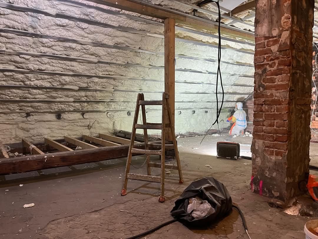 Poddasze po ociepleniu pianką PUR, widoczna warstwa izolacji na ścianach i stropie, drabina i pracownik w kombinezonie ochronnym w tle. Ceglany filar po prawej.