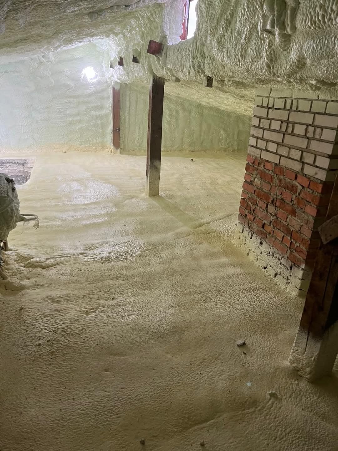 Poddasze po ociepleniu pianką PUR: widoczne drewniane elementy konstrukcyjne i fragment ściany z cegły, pokryte grubą warstwą jasnej piany izolacyjnej.