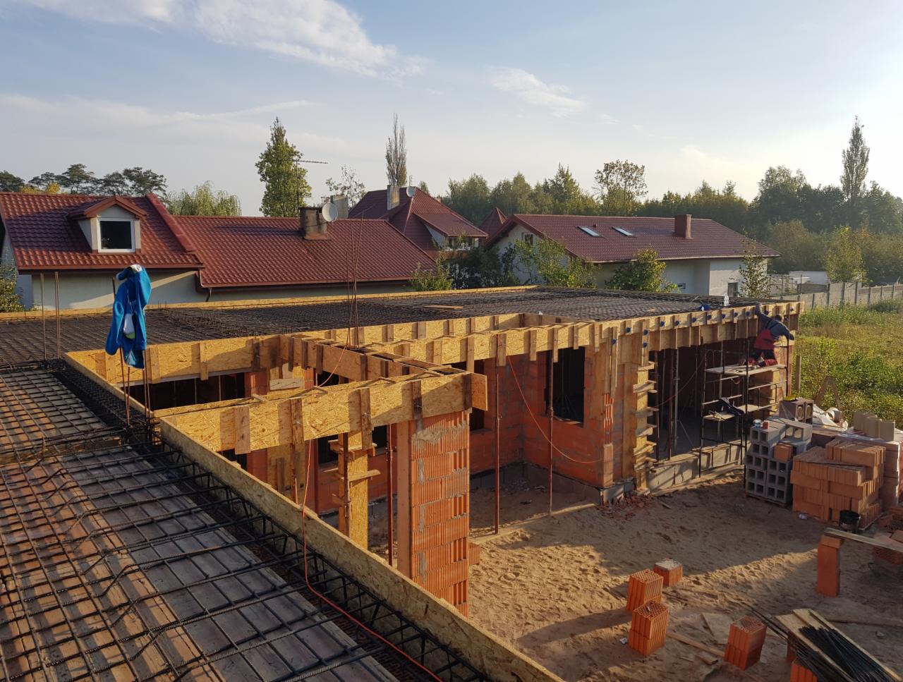 Budowa domu jednorodzinnego w trakcie wznoszenia ścian z cegły, widoczne drewniane szalunki i zbrojenie stropu, na drugim planie zabudowa mieszkalna i drzewa.