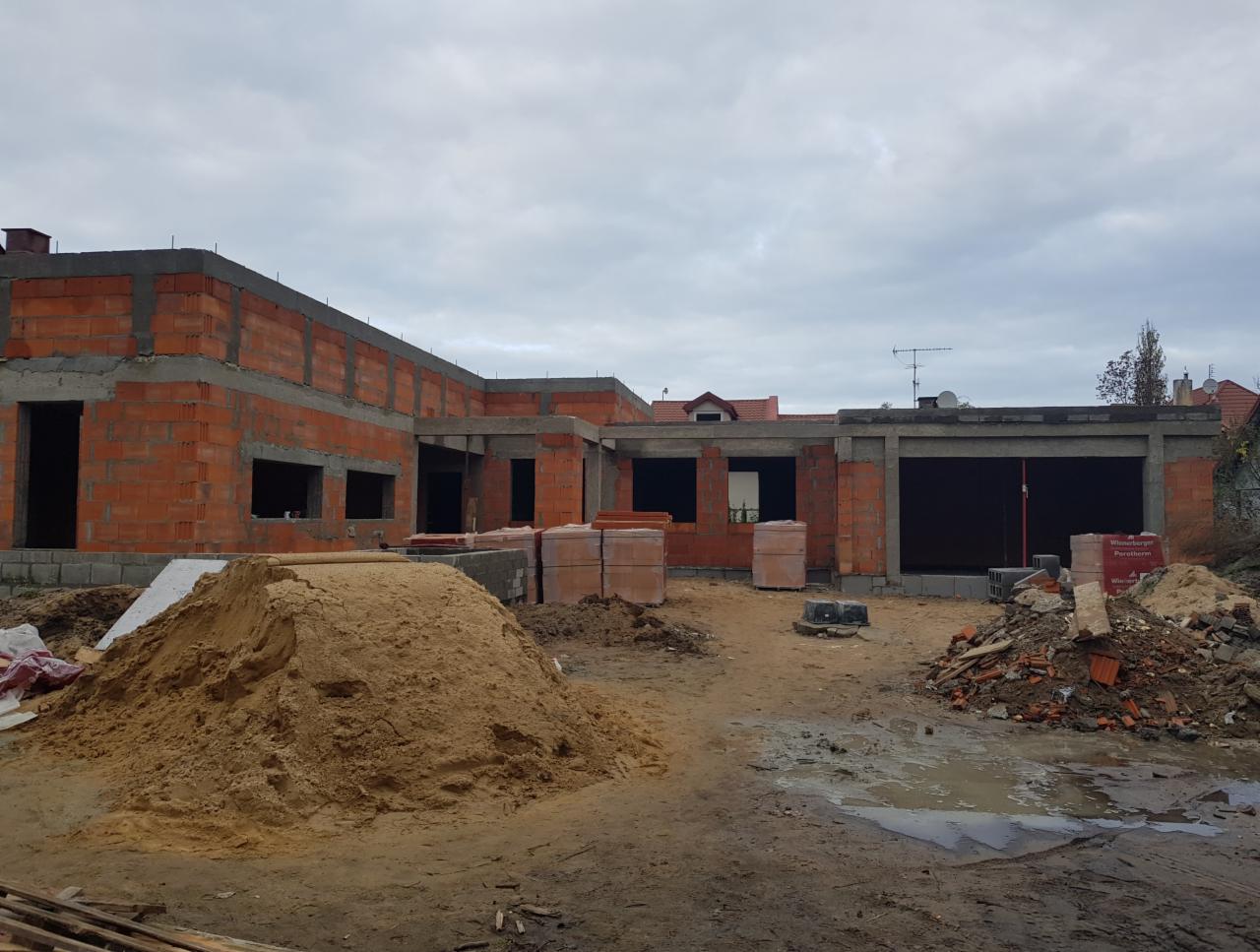 Wznoszenie ścian parterowego domu z czerwonej cegły ceramicznej, widoczne otwory okienne i drzwiowe, sterta piasku na pierwszym planie, pochmurne niebo w tle.