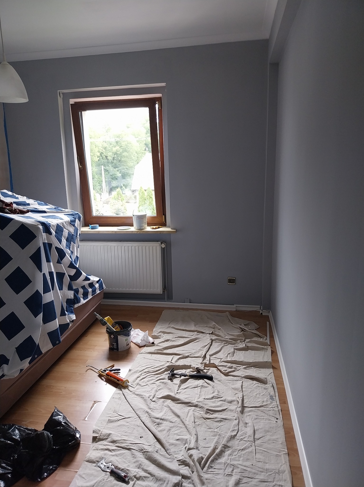 Pokój po malowaniu na szaro, zabezpieczone meble, widoczne narzędzia malarskie i wiadro z farbą. Okno z widokiem na zieleń. Świeżo pomalowane ściany.