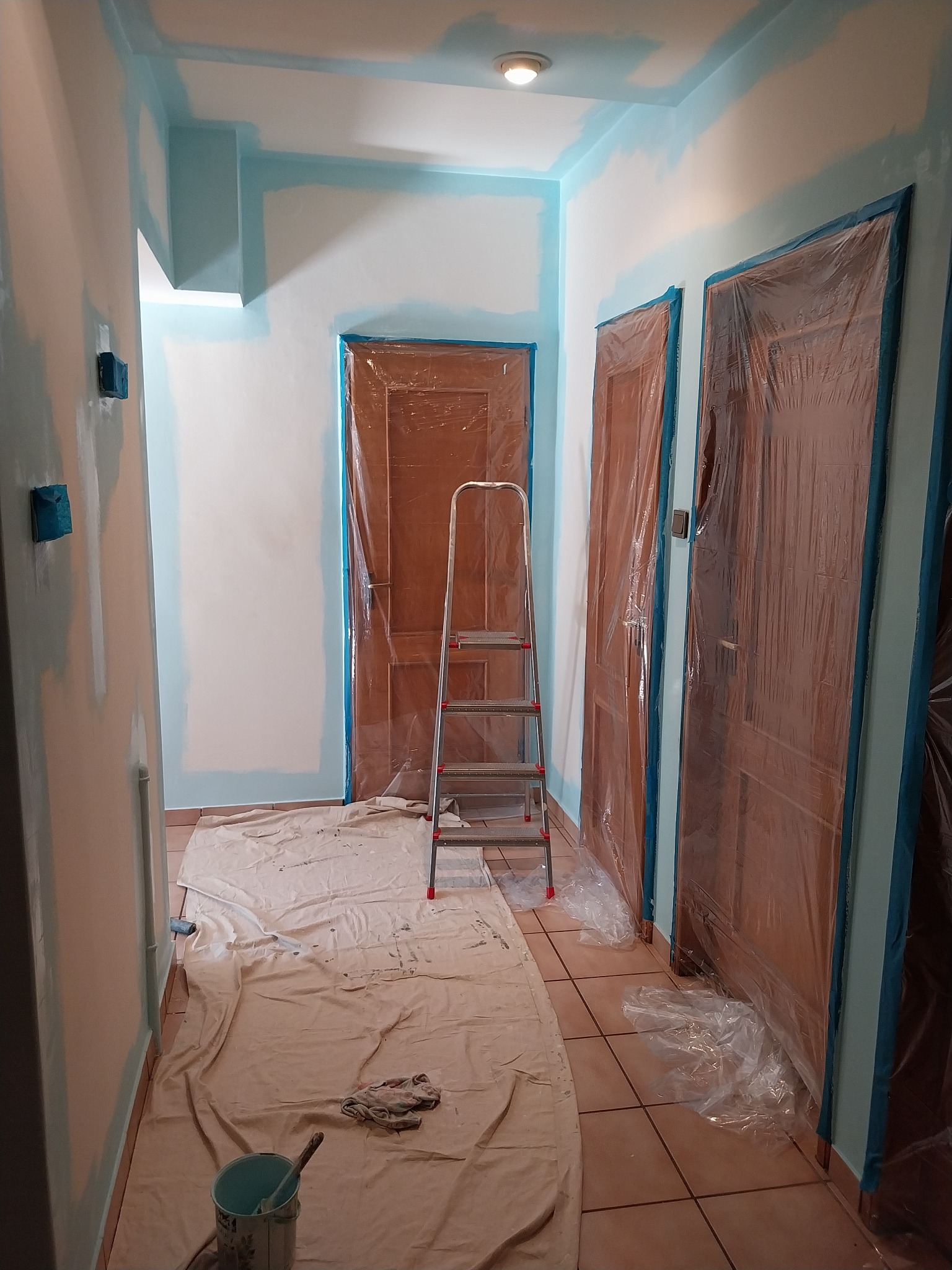 Korytarz w trakcie malowania na biało z niebieskimi akcentami. Drzwi zabezpieczone folią, widoczna drabina i akcesoria malarskie, podłoga zakryta płachtą.