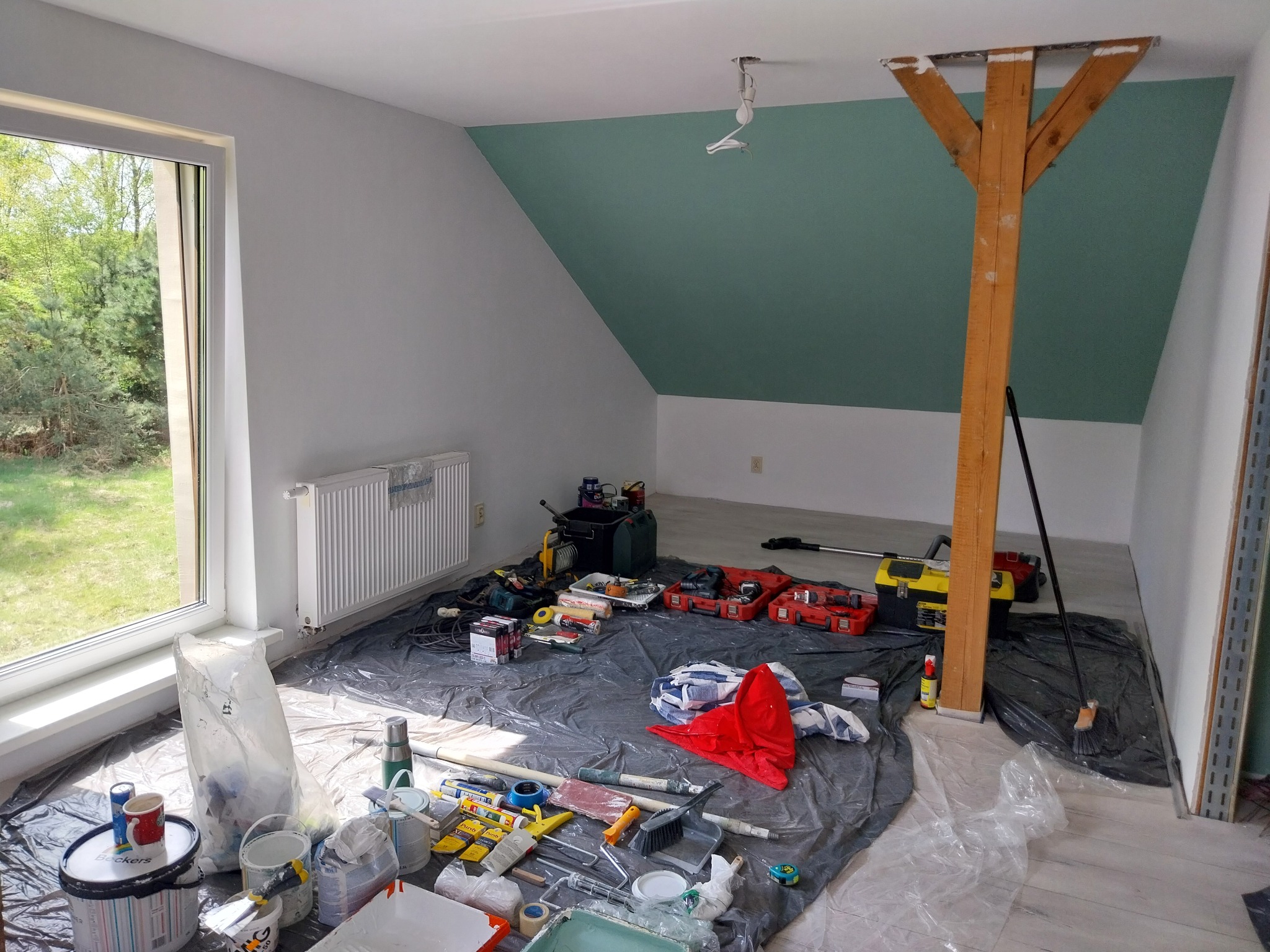 Malowanie poddasza w Ziębicach: ściany pomalowane na szaro i zielono, drewniany słup, okno, grzejnik i liczne narzędzia malarskie rozłożone na folii ochronnej.