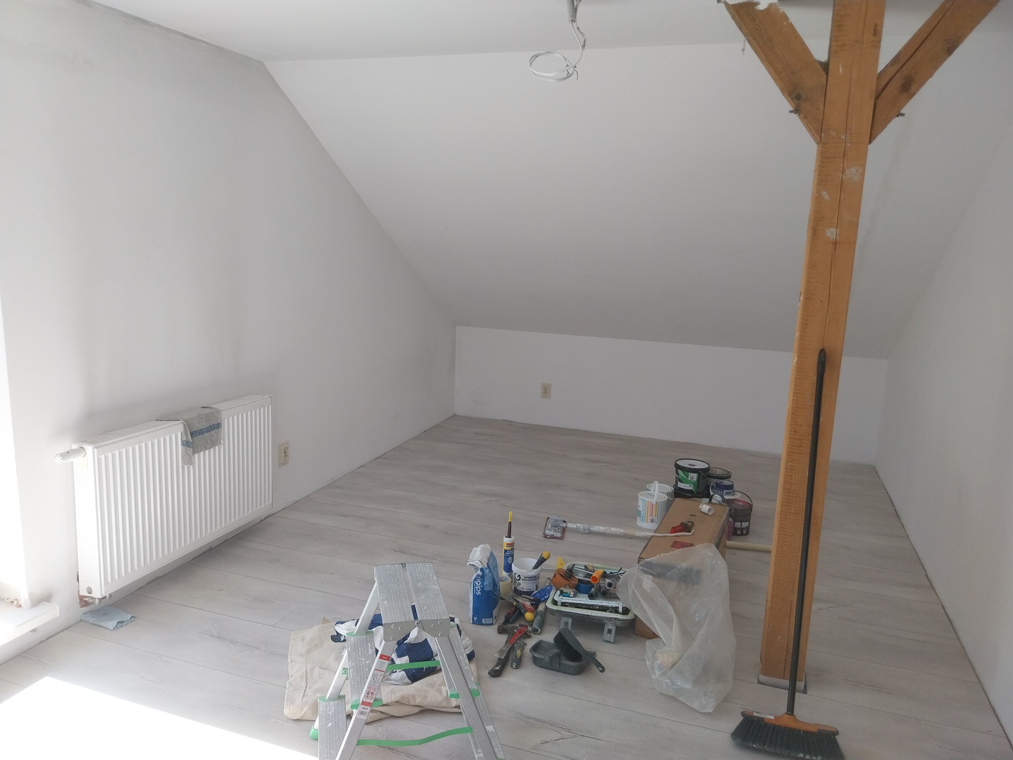 Pokój na poddaszu po malowaniu na biało, z widocznymi narzędziami malarskimi, drabiną i drewnianą belką konstrukcyjną. Świeżo pomalowane ściany i sufit.