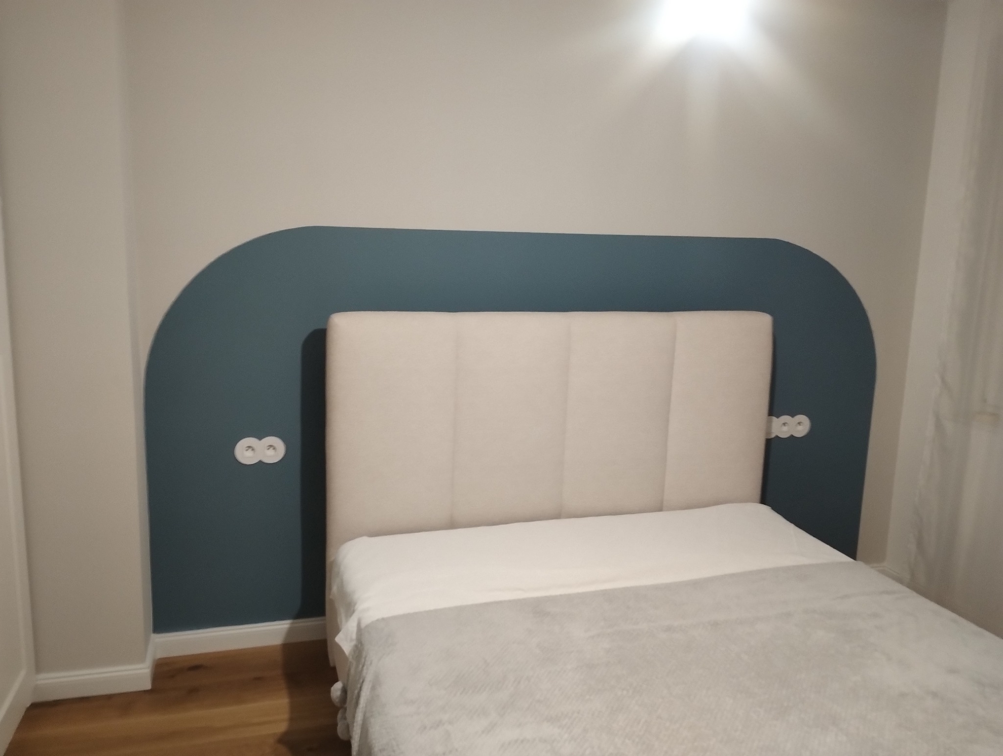 Sypialnia z łóżkiem i dekoracyjną, łukową ścianą w kolorze petrol, z dwoma gniazdkami elektrycznymi. Minimalistyczny design, jasne kolory, drewniana podłoga.