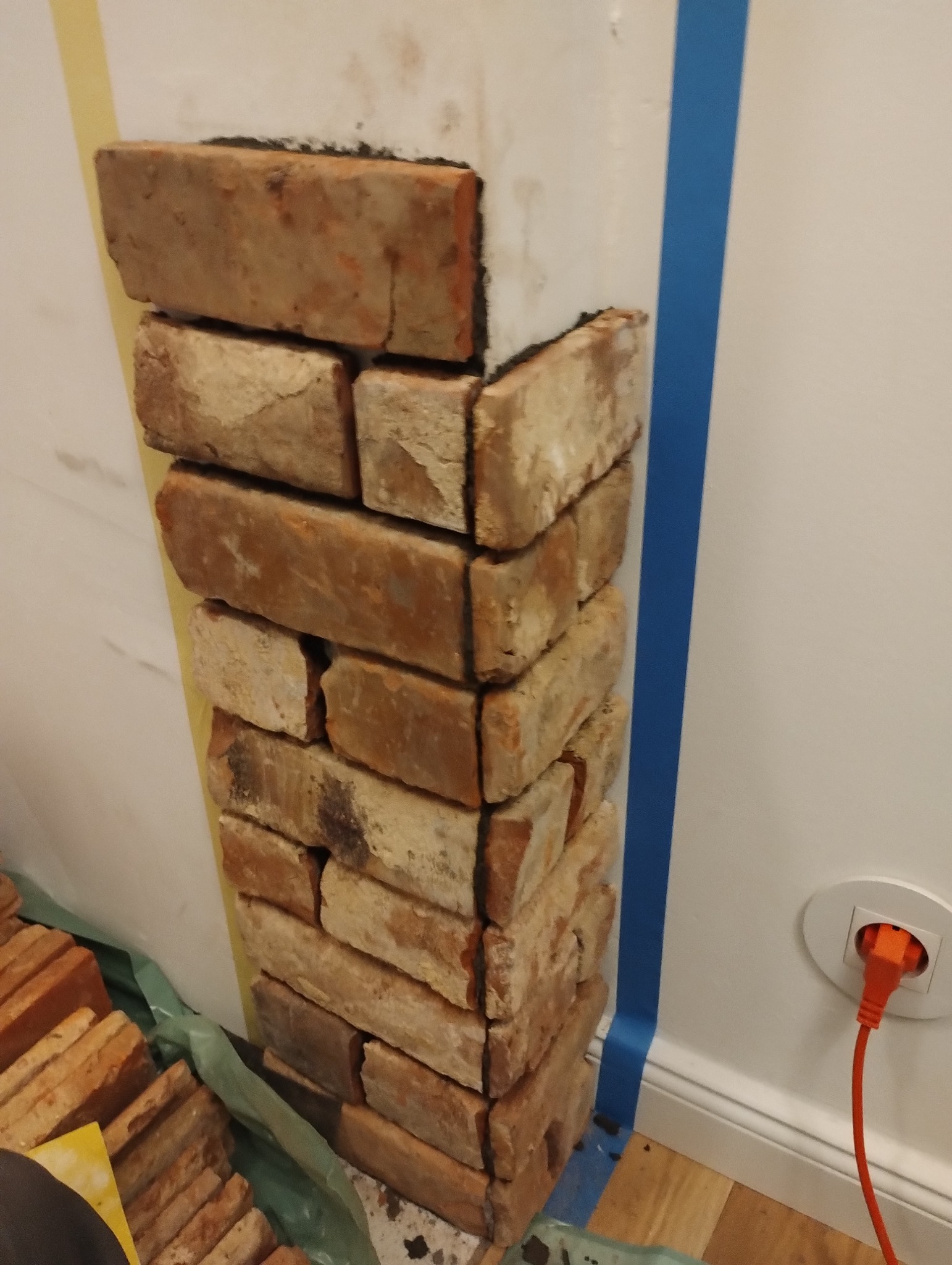 Ściana w trakcie dekorowania cegłą klinkierową, widoczne taśmy malarskie, przygotowane cegły i gniazdko elektryczne z wpiętym kablem.