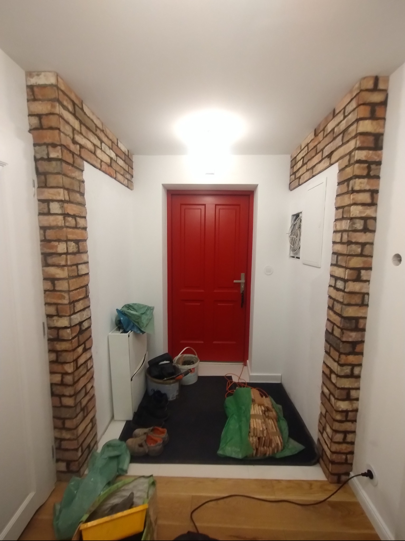 Korytarz z dekoracyjnymi filarami z cegły po bokach wejścia. Czerwone drzwi na wprost, podłoga wyłożona czarną wykładziną, widoczne ślady prac wykończeniowych.