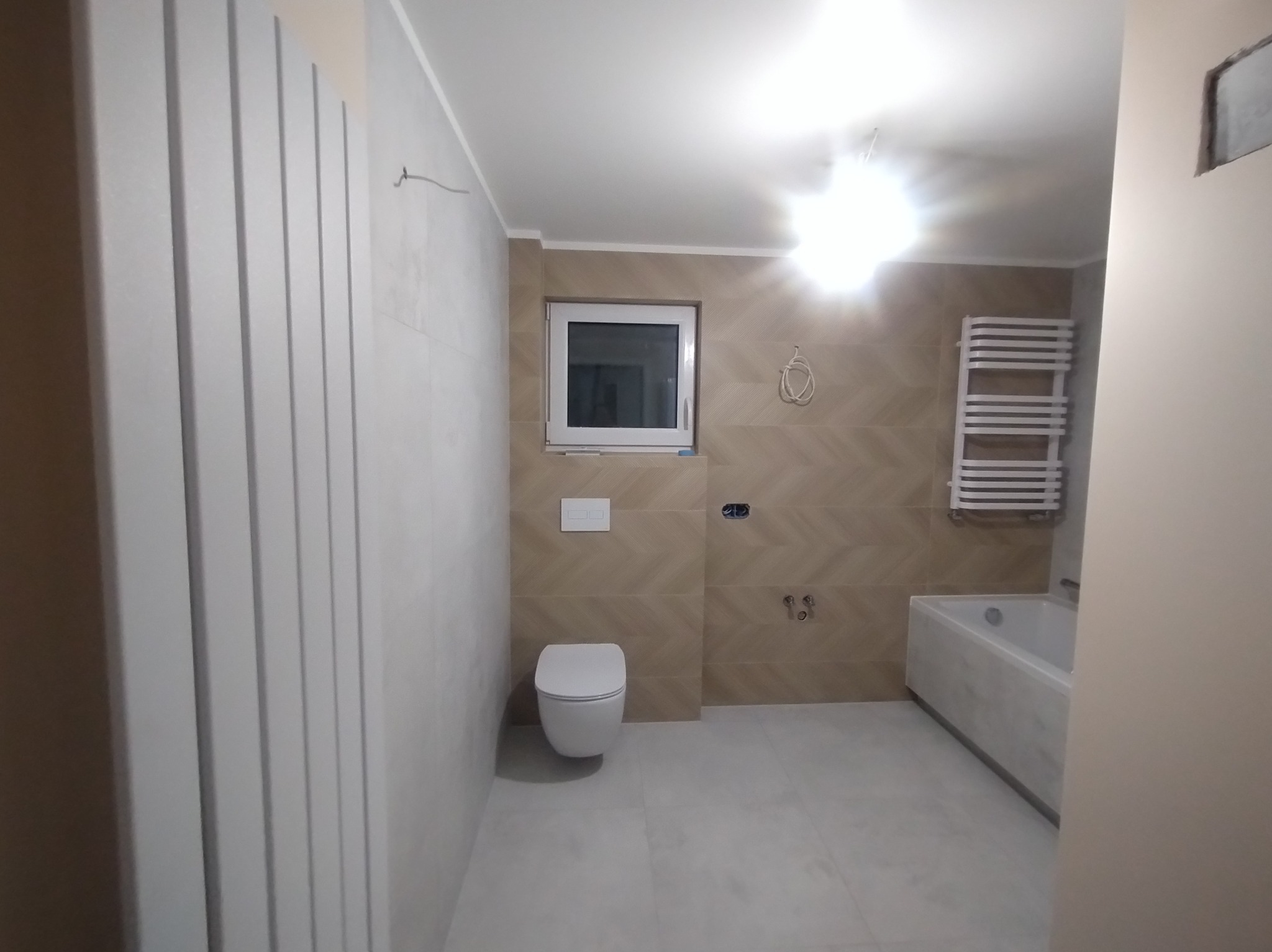 Jasna łazienka po remoncie. Widoczna toaleta podwieszana, grzejnik drabinkowy, wanna, okno. Na ścianach płytki w odcieniach beżu i bieli. Minimalistyczny styl.