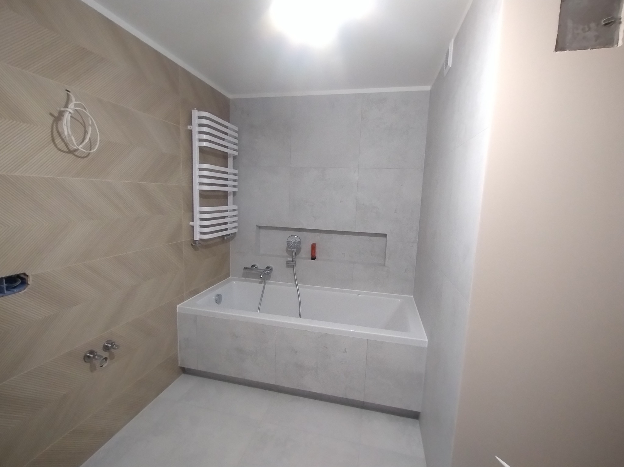 Wykończona łazienka z wanną obudowaną płytkami, grzejnikiem drabinkowym i dekoracyjnymi płytkami w jodełkę na ścianie. Widoczne przyłącza wodne i oświetlenie.