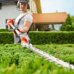 FACHOWIEC PODLASIE - Mężczyzna w kombinezonie przycina żywopłot elektrycznymi nożycami, w tle dom z czerwoną dachówką i błękitne niebo.