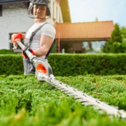 Mężczyzna w kombinezonie przycina żywopłot elektrycznymi nożycami, w tle dom z czerwoną dachówką i błękitne niebo.