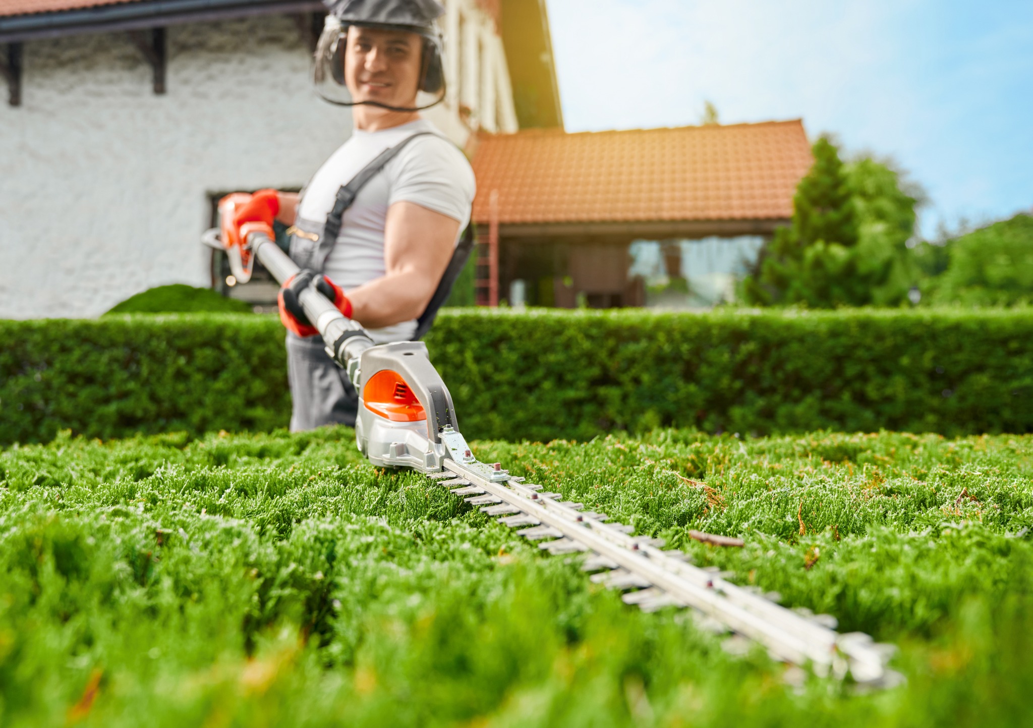 Mężczyzna w kombinezonie przycina żywopłot elektrycznymi nożycami, w tle dom z czerwoną dachówką i błękitne niebo.