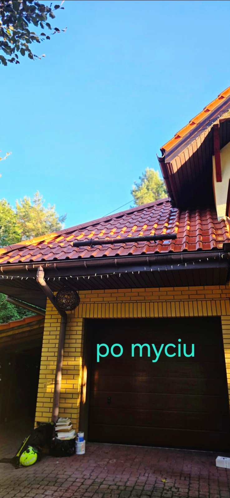 Czysty dach z czerwonej dachówki po umyciu, widoczny fragment elewacji budynku z żółtej cegły i brama garażowa. Na dole leżą narzędzia i materiały.