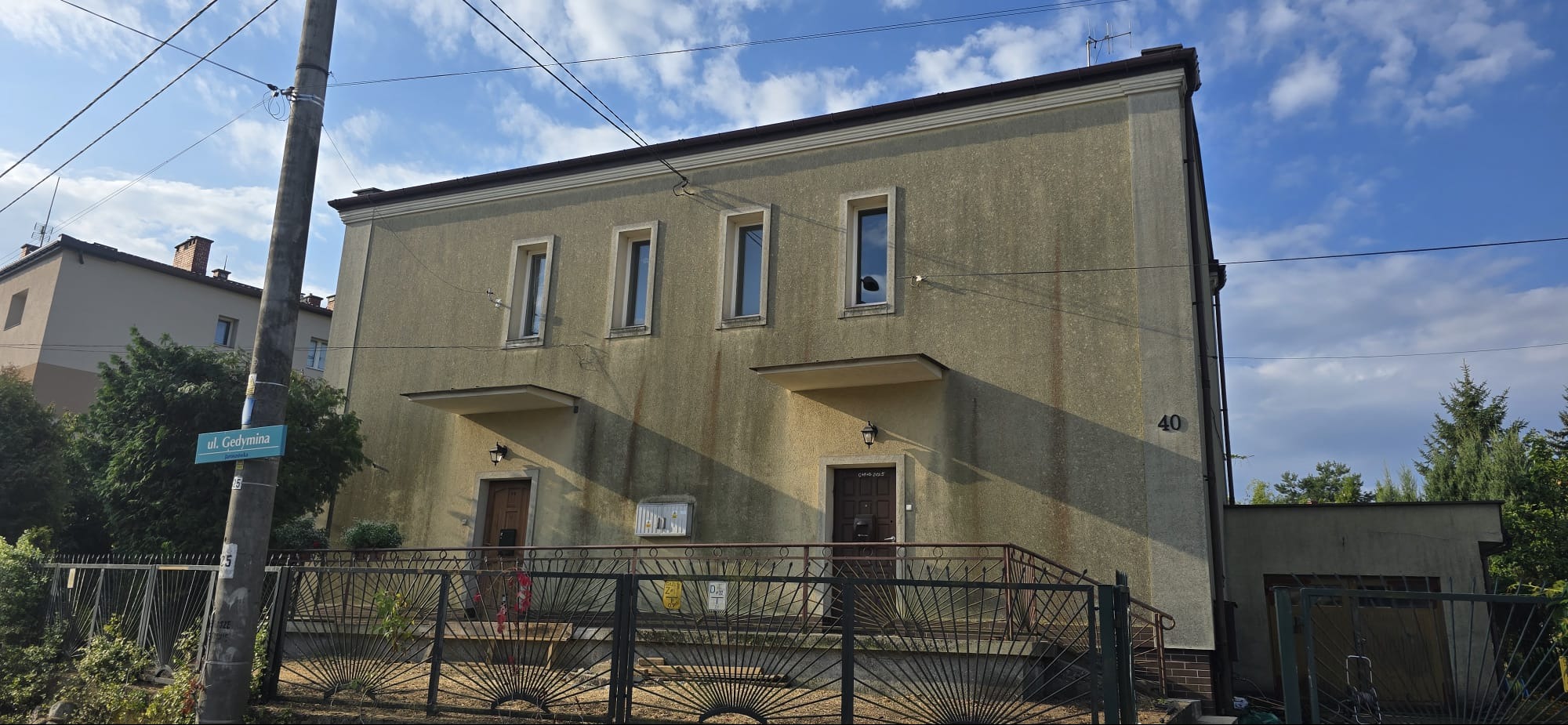 Dwukondygnacyjny budynek z szarą elewacją, widoczny z ulicy Gedymina, z metalowym ogrodzeniem i numerem 40, oświetlony naturalnym światłem słonecznym.