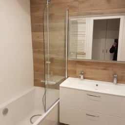 USŁUGI BUDOWLANE MIROSŁAW SIEDLECKI - Nowoczesna łazienka po remoncie w Kolbuszowej: biała armatura, kabina prysznicowa ze szklanym panelem, drewnopodobne płytki na ścianie i lustro z szafką.