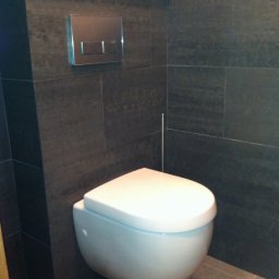 USŁUGI BUDOWLANE MIROSŁAW SIEDLECKI - Nowoczesna toaleta podwieszana z chromowanym przyciskiem spłukującym na tle ciemnych, grafitowych płytek w wykończonej łazience.