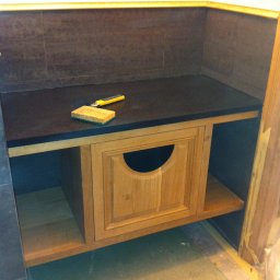 USŁUGI BUDOWLANE MIROSŁAW SIEDLECKI - Wykończona wnęka w łazience z drewnianą szafką z okrągłym otworem. Na blacie leży gąbka i nożyk. Tło stanowią ciemne płytki i biała ściana.