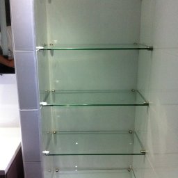 USŁUGI BUDOWLANE MIROSŁAW SIEDLECKI - Szklane półki wbudowane w ścianę, wsporniki w kolorze srebrnym. Widoczne płytki. Minimalistyczny design, oświetlenie punktowe na górze.