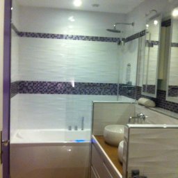 USŁUGI BUDOWLANE MIROSŁAW SIEDLECKI - Nowoczesna łazienka z białą wanną, mozaiką i umywalką nablatową. Widoczne drewniane panele podłogowe i szklana kabina prysznicowa. Oświetlenie sufitowe.