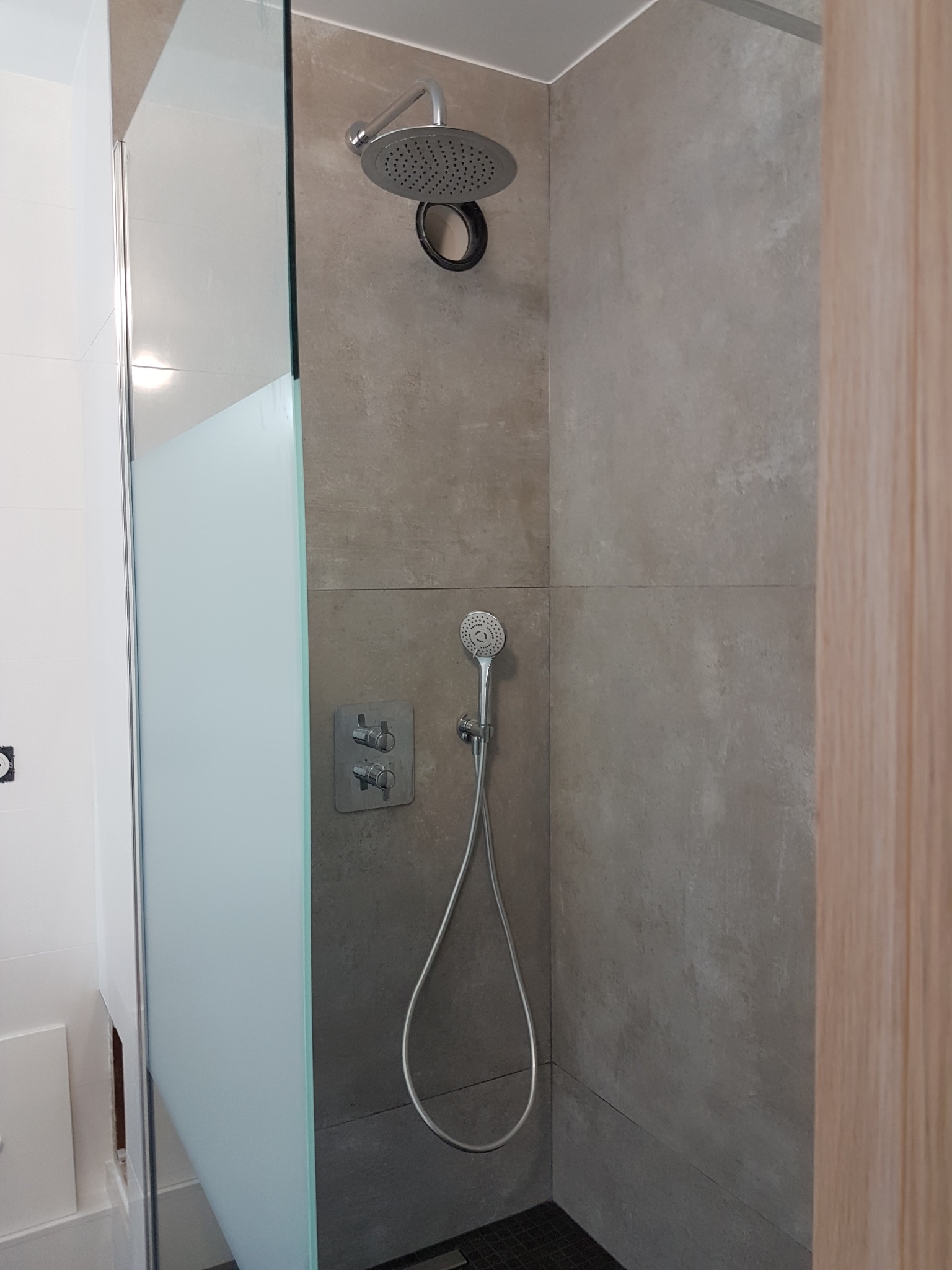 Nowoczesna kabina prysznicowa z deszczownicą i szklaną ścianką, wykończona szarymi płytkami. Minimalistyczny design, widoczne detale armatury i odpływ liniowy.