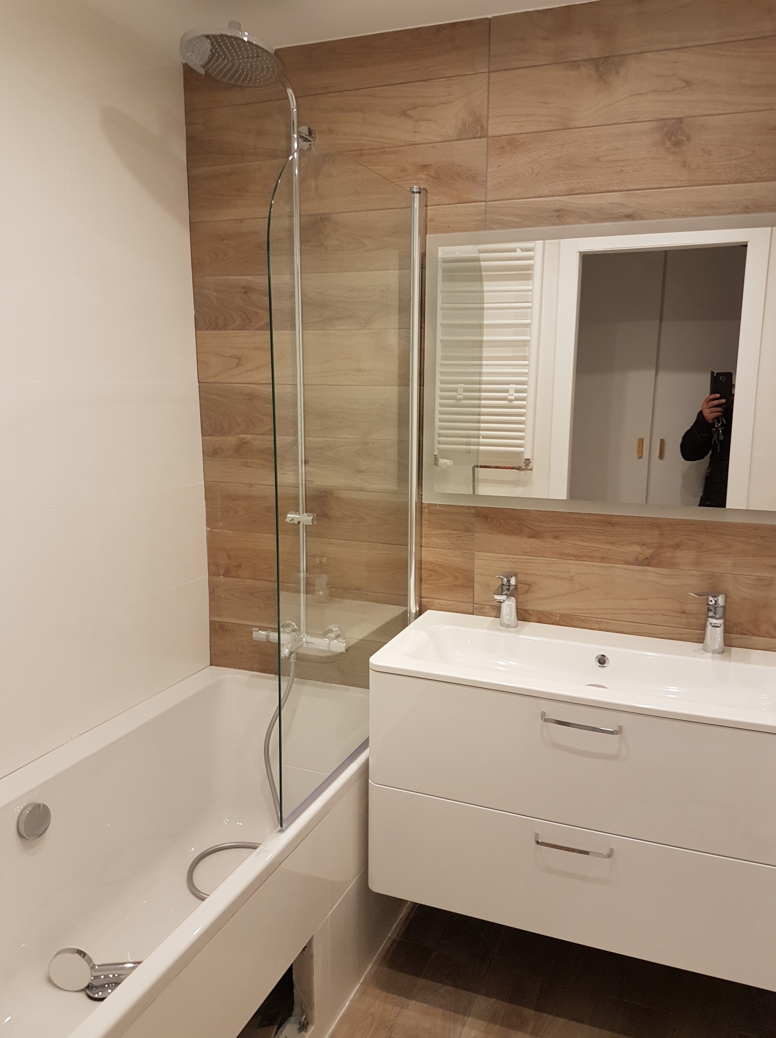 Nowoczesna łazienka po remoncie w Kolbuszowej: biała armatura, kabina prysznicowa ze szklanym panelem, drewnopodobne płytki na ścianie i lustro z szafką.