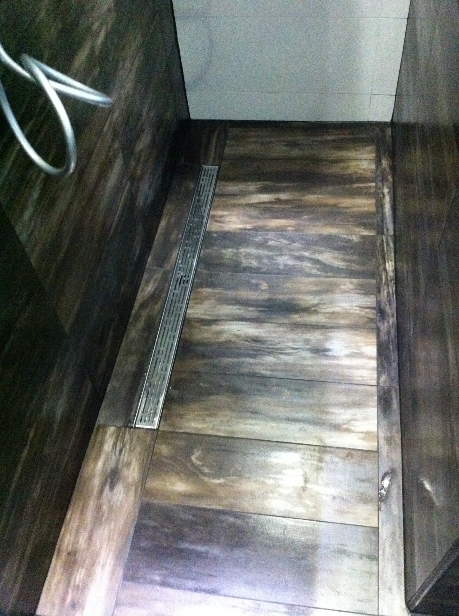 Ukończona łazienka: podłoga i ściany wyłożone płytkami imitującymi drewno, z liniowym odpływem prysznicowym w kolorze stali nierdzewnej.