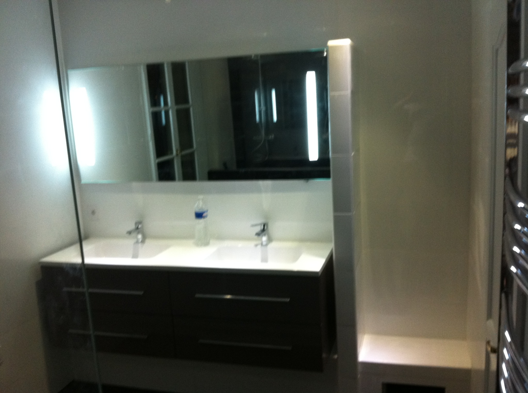 Nowoczesna łazienka z podwójną umywalką, dużym lustrem z podświetleniem LED i ciemną szafką. Widoczne płytki na ścianach i fragment grzejnika drabinkowego.