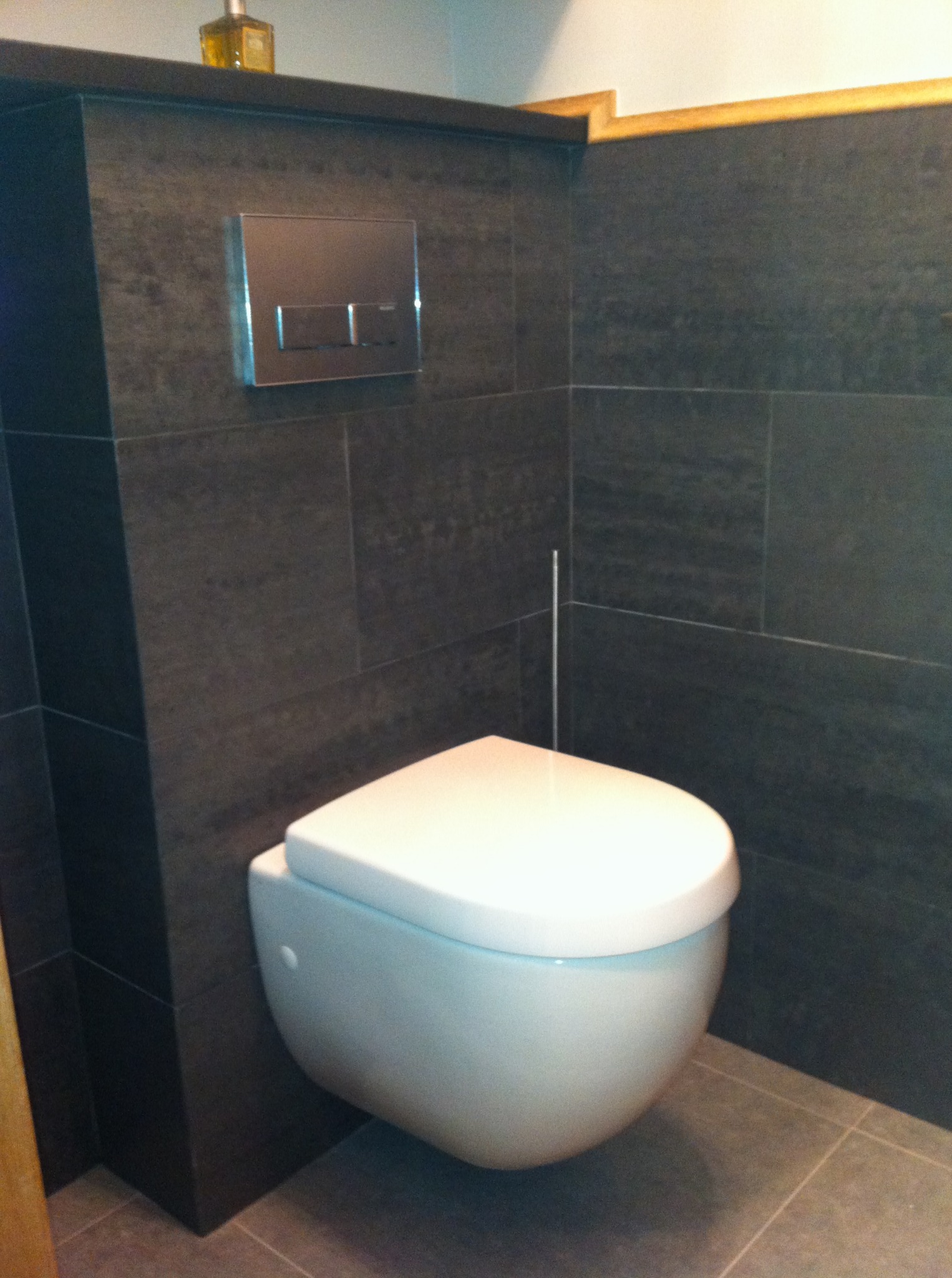 Nowoczesna toaleta podwieszana z chromowanym przyciskiem spłukującym na tle ciemnych, grafitowych płytek w wykończonej łazience.