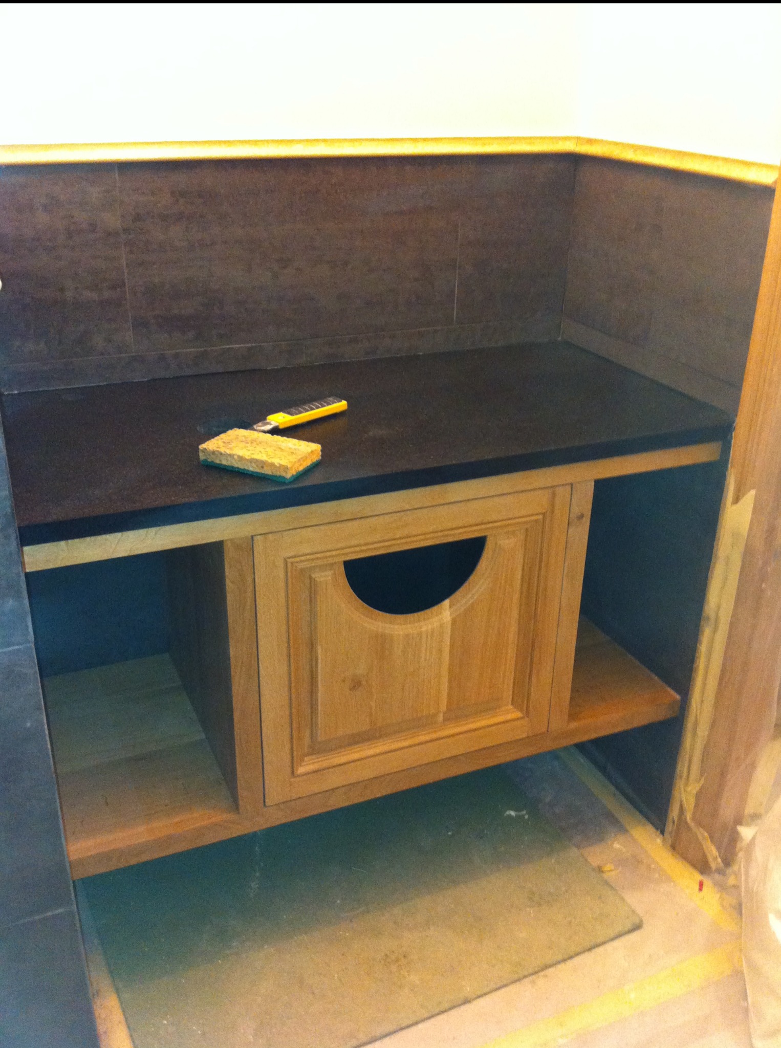 Wykończona wnęka w łazience z drewnianą szafką z okrągłym otworem. Na blacie leży gąbka i nożyk. Tło stanowią ciemne płytki i biała ściana.