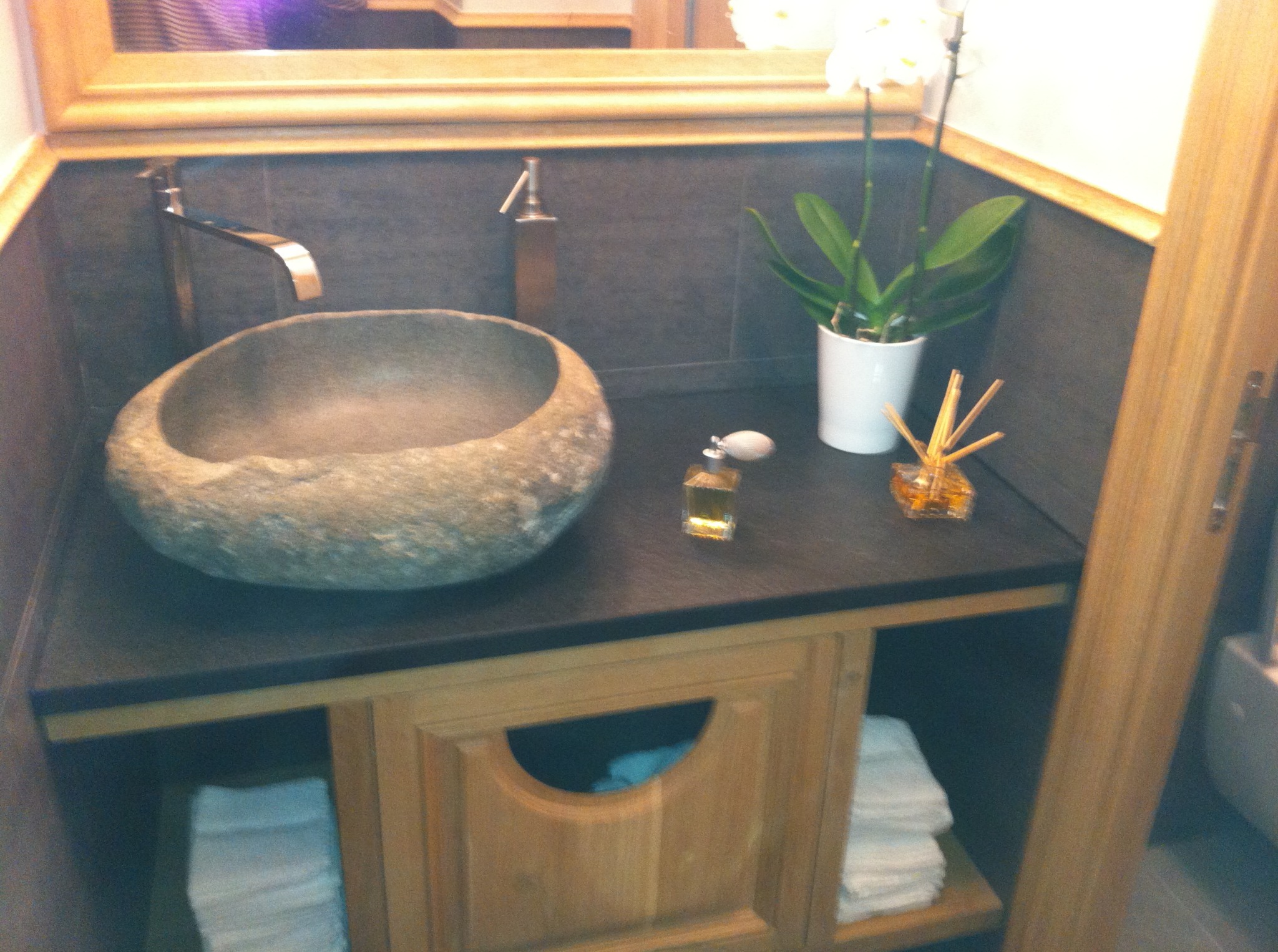 Stylowa łazienka: kamienna umywalka w kształcie misy, chromowana bateria, dozownik mydła i dekoracje na blacie z ciemnego kamienia. Pod umywalką drewniana szafka z ręcznikami.