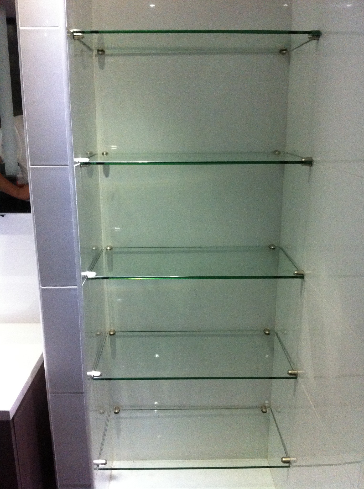 Szklane półki wbudowane w ścianę, wsporniki w kolorze srebrnym. Widoczne płytki. Minimalistyczny design, oświetlenie punktowe na górze.