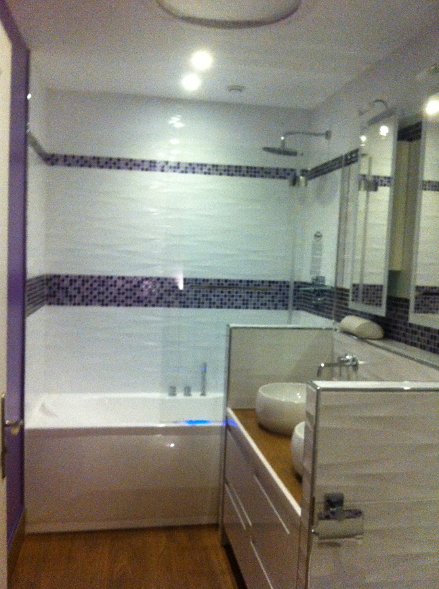 Nowoczesna łazienka z białą wanną, mozaiką i umywalką nablatową. Widoczne drewniane panele podłogowe i szklana kabina prysznicowa. Oświetlenie sufitowe.