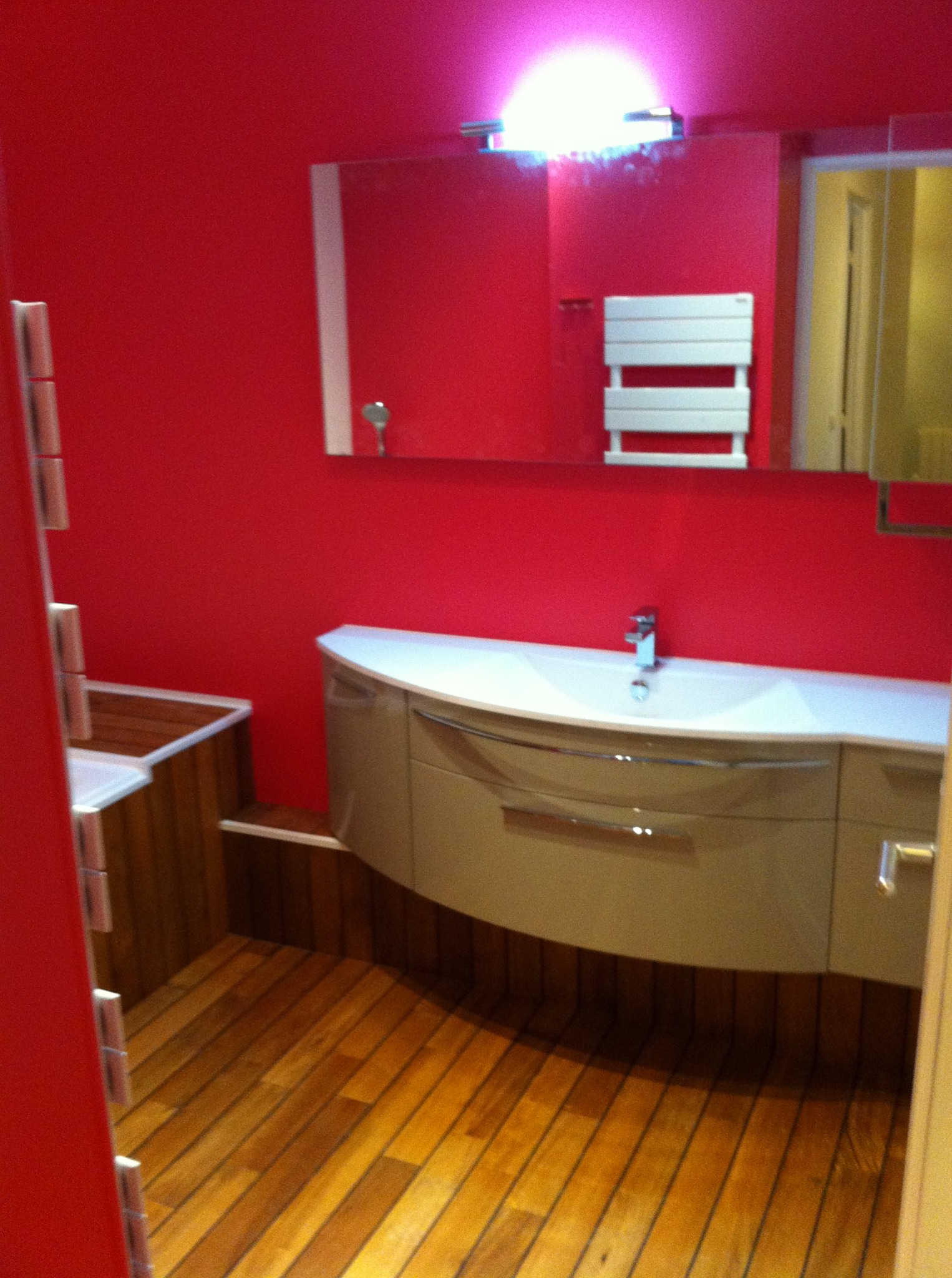 Czerwona łazienka z drewnianą podłogą, umywalką z szafką i grzejnikiem na ścianie. Lustro z oświetleniem nad umywalką. Nietypowe wykończenie w drewnie.