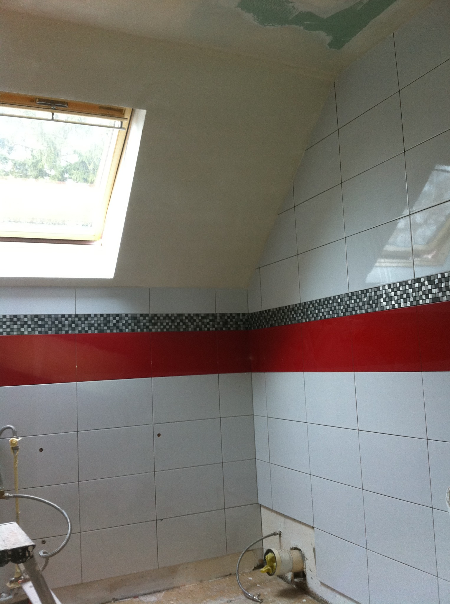 Ściana łazienki po remoncie z białymi płytkami, czerwoną wstawką i mozaiką, okno dachowe. Widoczne niedokończone podłączenia hydrauliczne.