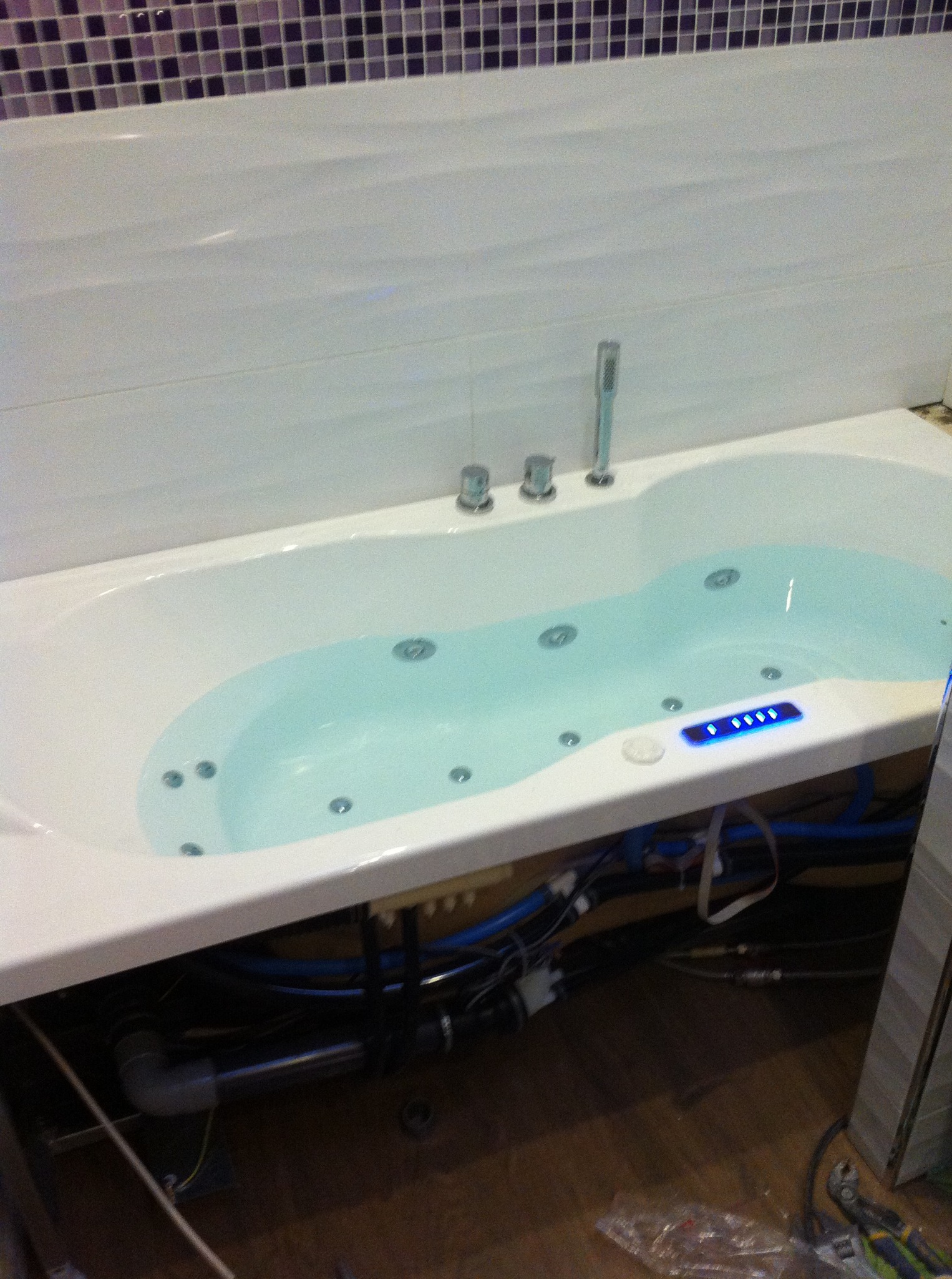 Biała wanna z hydromasażem wypełniona wodą, widoczne rury i instalacja pod wanną, oraz fragment mozaiki na ścianie.