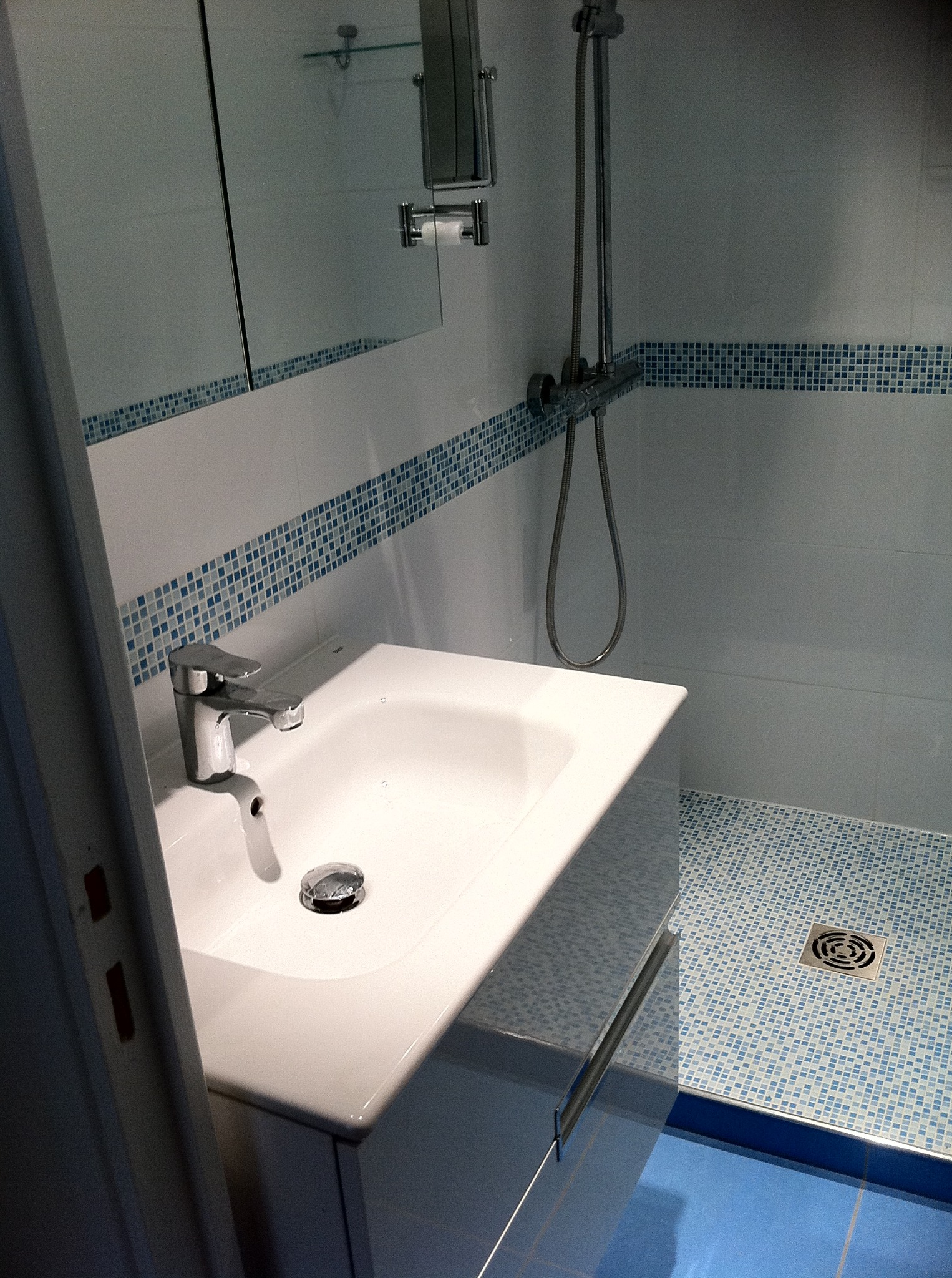 Biała umywalka z chromowaną baterią w wykończonej łazience z niebieską mozaiką. Widoczny kabina prysznicowa z odpływem. Częściowo widoczne drzwi.