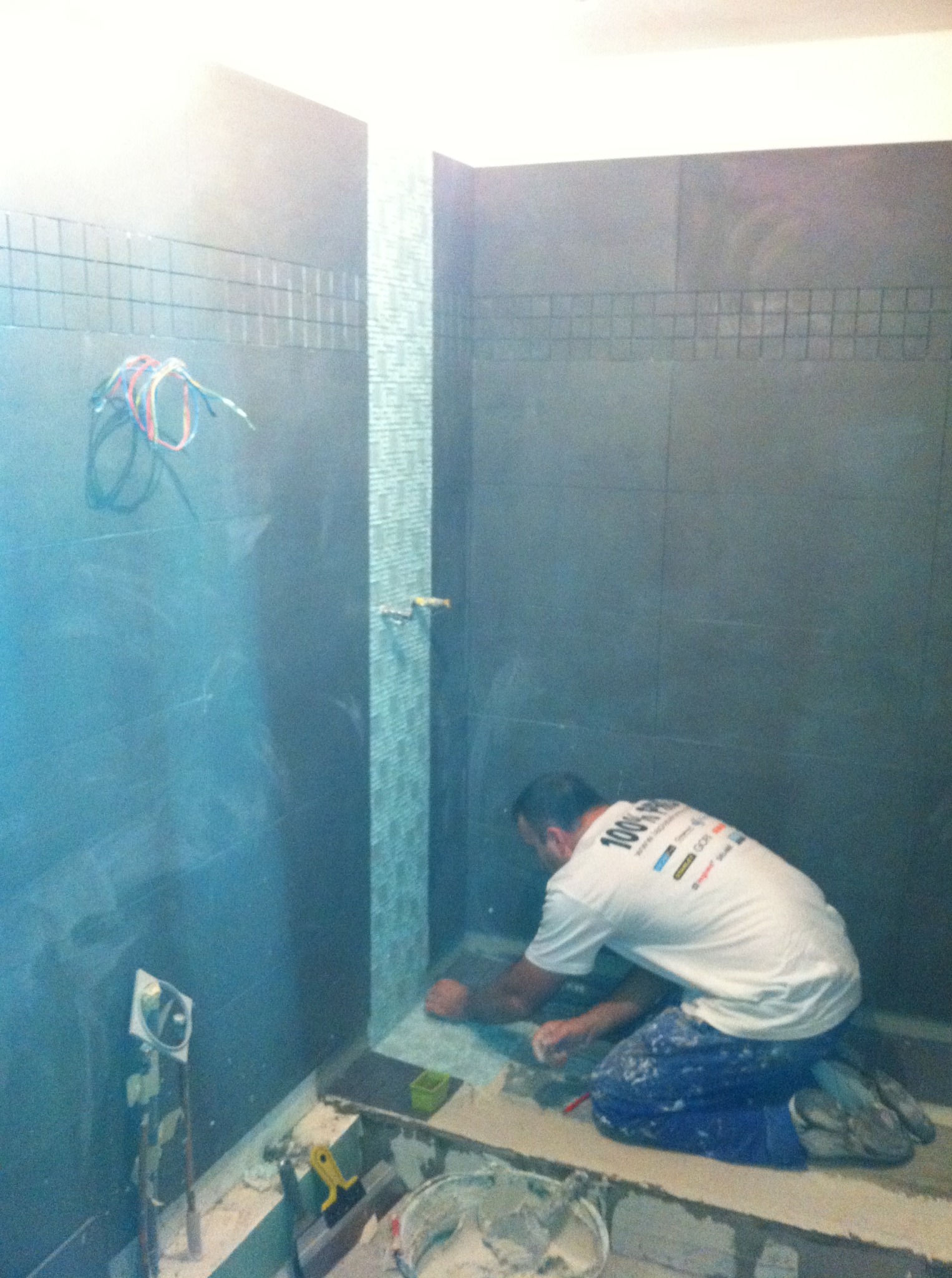 Układanie mozaiki w kabinie prysznicowej. Glazurnik na kolanach precyzyjnie dociska małe płytki. Widoczne narzędzia i klej. Szare, duże płytki na ścianach.