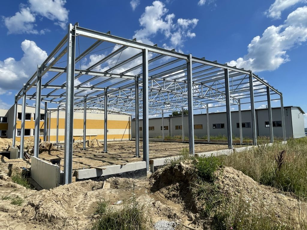 Stalowa konstrukcja hali w trakcie budowy, widoczne fundamenty i słupy, w tle budynek z żółtymi pasami, błękitne niebo z chmurami.