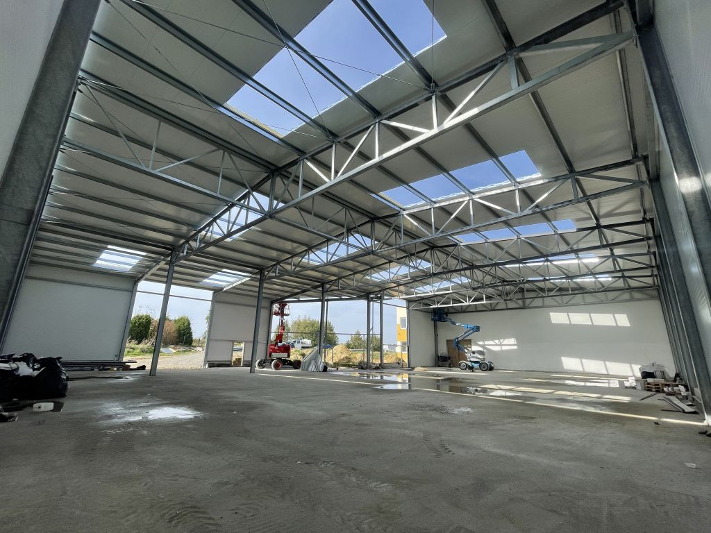 Wnętrze hali w trakcie budowy z widoczną konstrukcją stalową, oświetleniem dachowym i sprzętem budowlanym w tle. Widoczne ślady po budowie na posadzce.