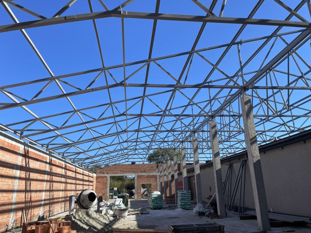 Konstrukcja stalowa hali w trakcie budowy, widok z dołu na szkielet dachu i ściany z cegły. Na dole widoczna betoniarka i materiały budowlane.