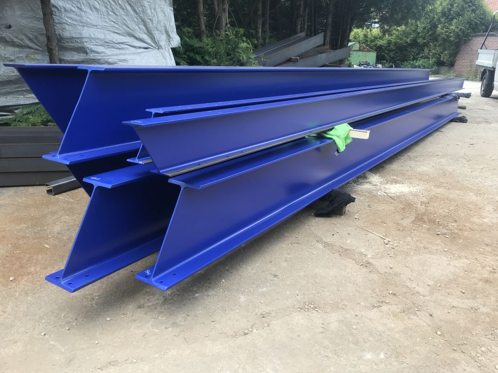 Stalowe, niebieskie belki H leżące na ziemi, przygotowane do budowy hali. Widoczne otwory montażowe i świeża farba. W tle zieleń drzew i część samochodu dostawczego.