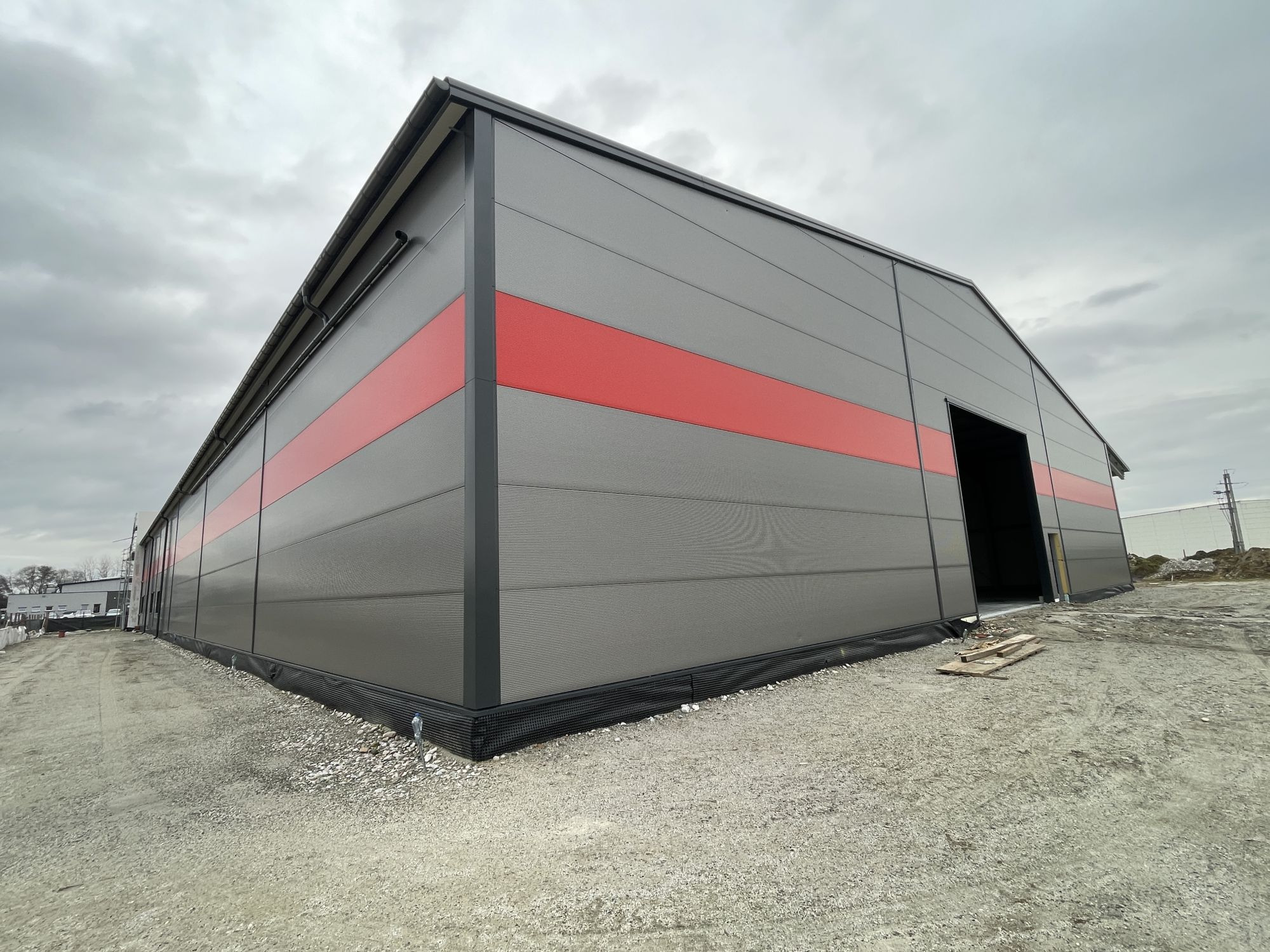 Szara hala z płyty warstwowej, z poziomym czerwonym pasem. Widoczny fragment dachu i otwarte wejście. Teren wokół hali przygotowany pod zagospodarowanie.
