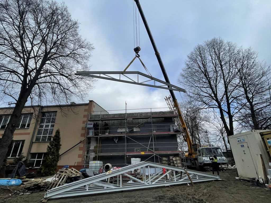 Budowa hali: dźwig podnosi stalową konstrukcję dachu na budynek w trakcie wznoszenia. Widoczne rusztowania i elementy konstrukcyjne na ziemi.